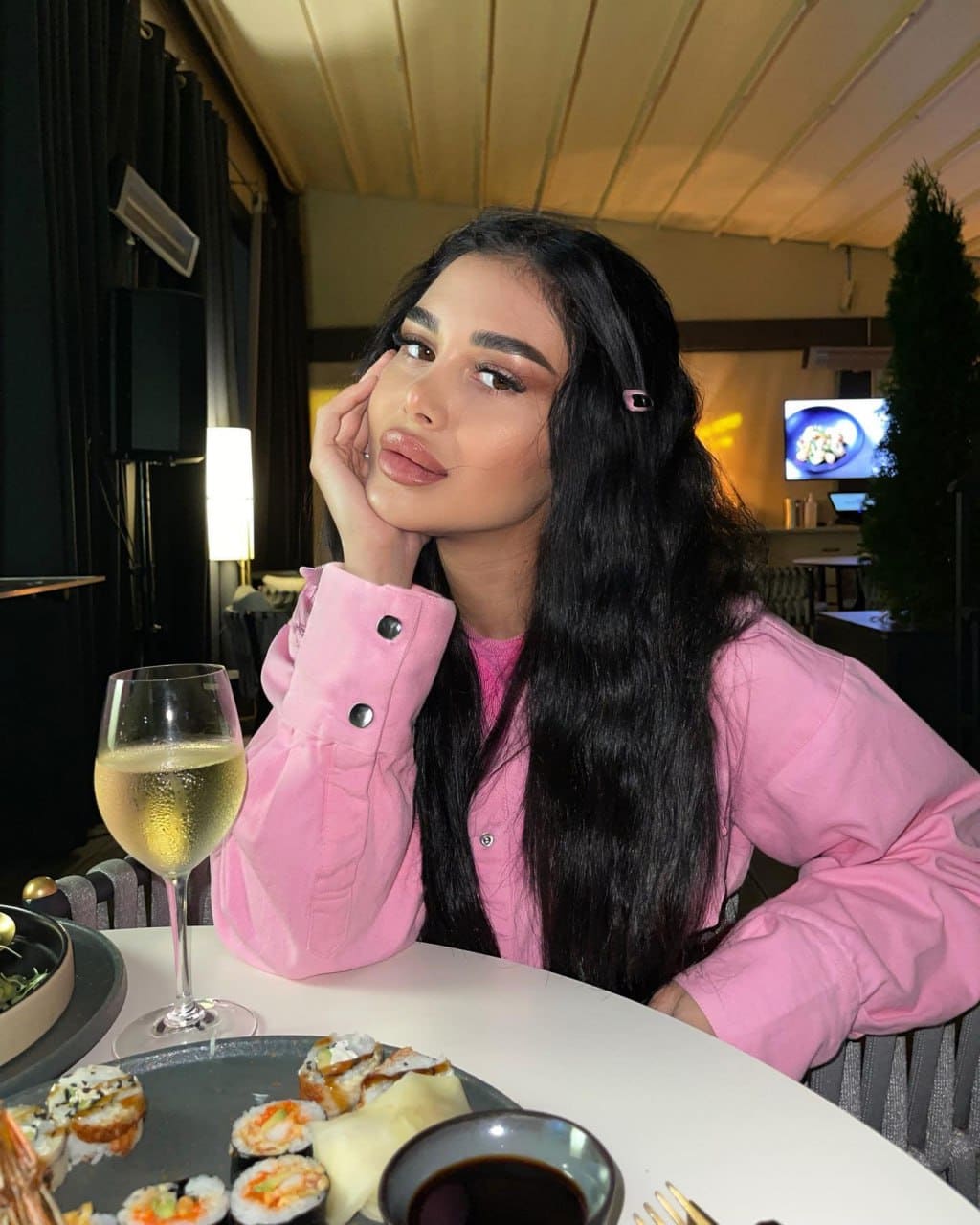 Sabina sits at a white restaurant table in a pink jacket, resting her chin on her hand beside a glass of white wine and a plate of sushi.