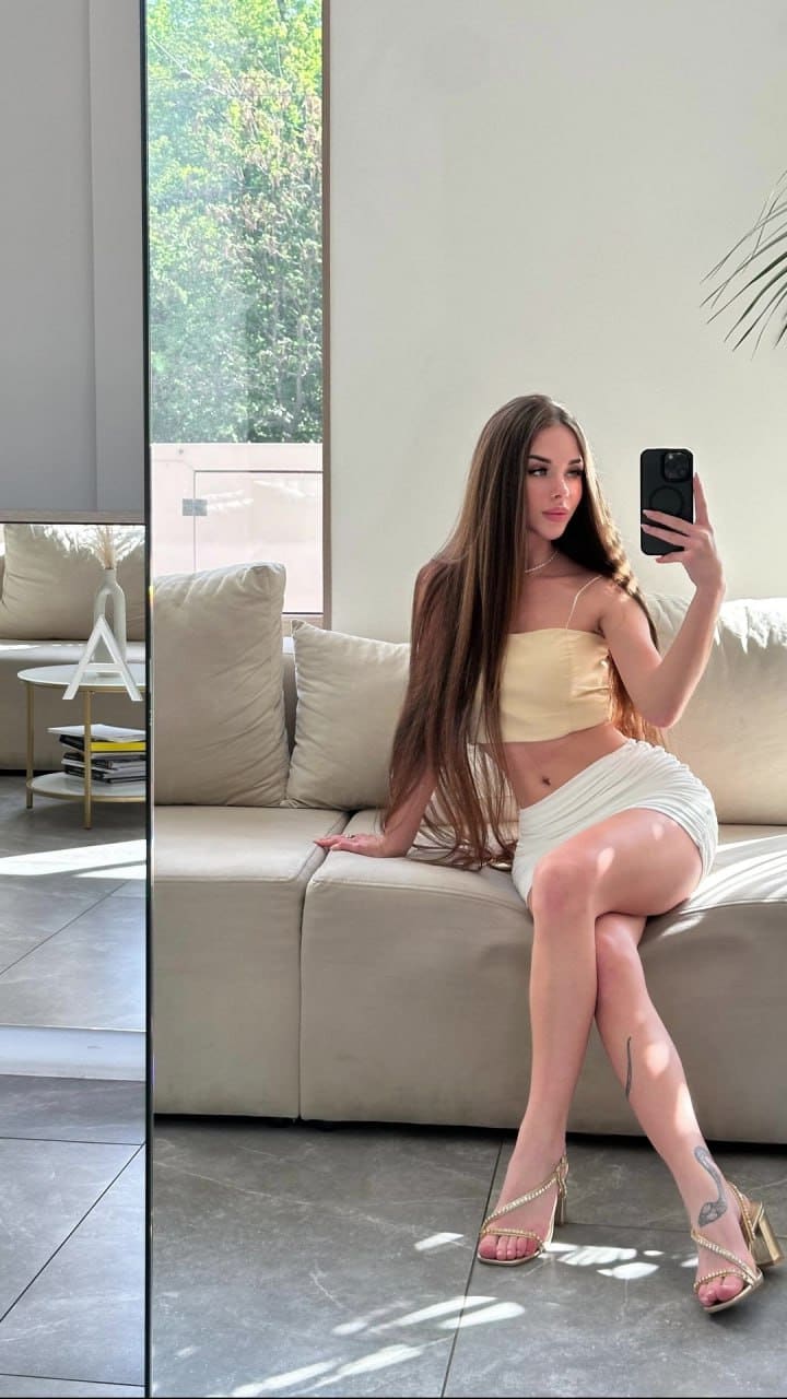 A woman in a yellow tube top and white skirt takes a mirror selfie on a beige sofa, wearing strappy high heels.