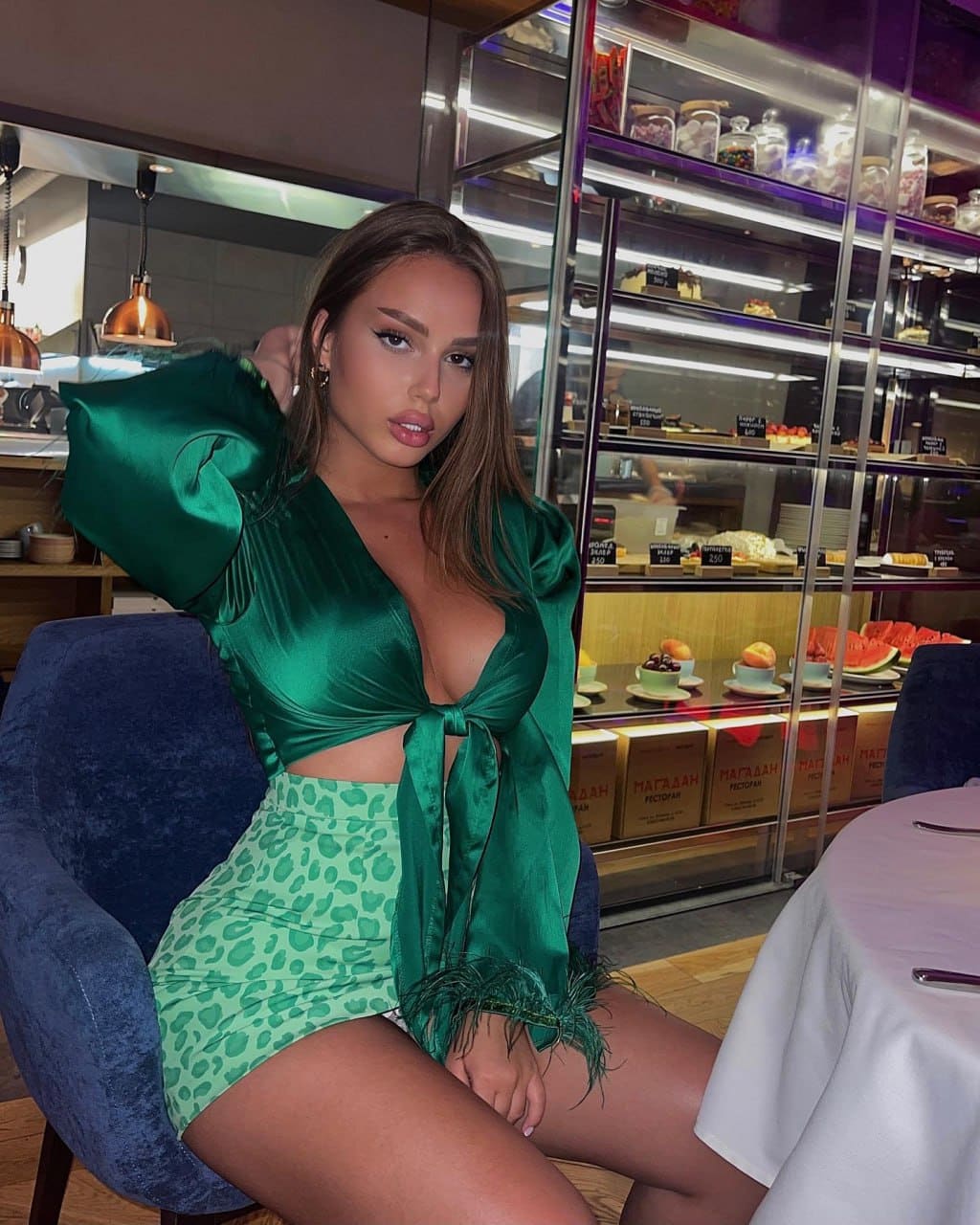 She sits on a blue chair in a cafe, wearing a green satin top and leopard-print skirt near a pastry case.