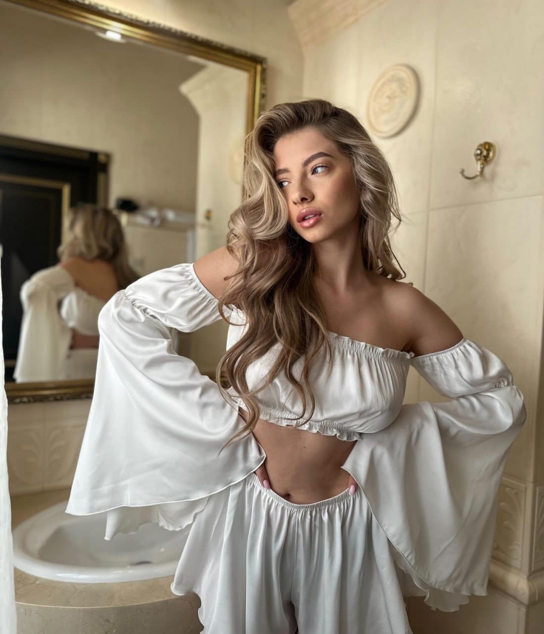 Posing in a tiled bathroom wearing a white off-shoulder crop top with bell sleeves and matching pants, hands on hips.