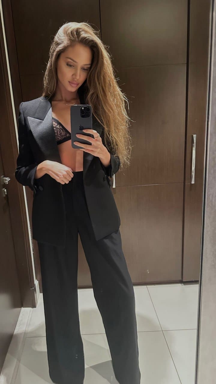 Blonde woman in an open black blazer and lace bra takes a mirror selfie, wearing wide-leg trousers in a room with dark wood paneling.