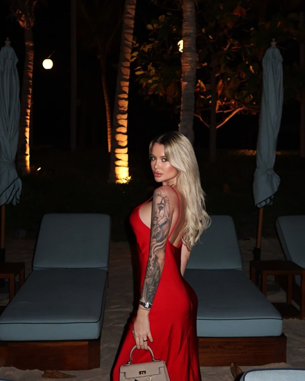 A woman with a full-sleeve tattoo stands in a red backless dress holding a handbag, with palm trees and lounge chairs behind her.