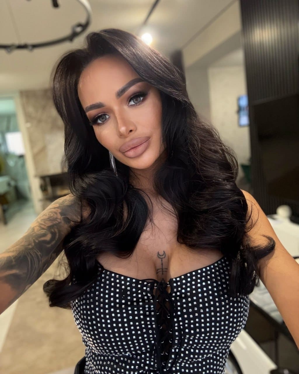 A woman with a tattooed arm poses in a black and white patterned lace-up top with bare shoulders in a modern indoor setting.
