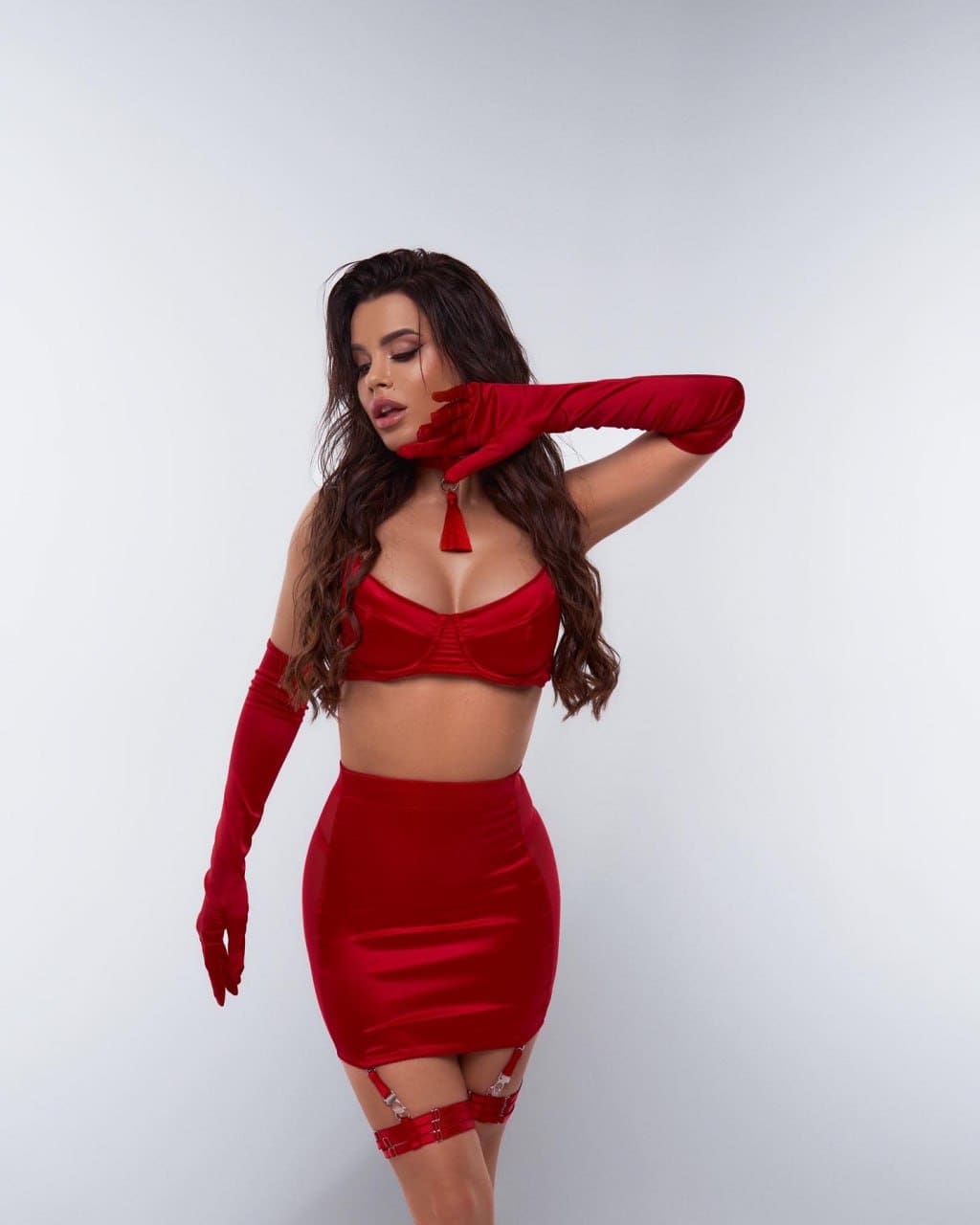 A woman poses against a plain background in a red satin bra, mini skirt, thigh-high garters, and long red gloves.