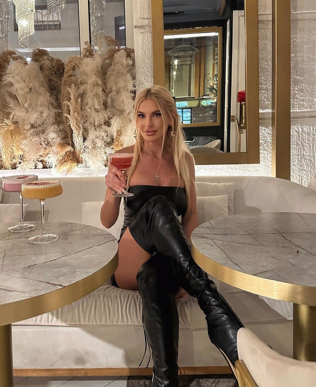 A blonde woman in a black mini dress and leather boots sits on a white sofa holding a cocktail.