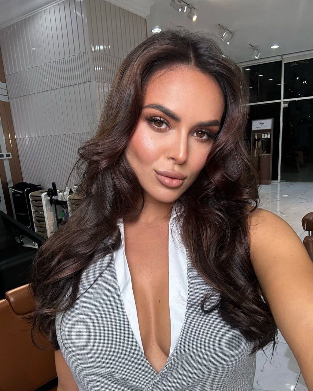 Milena poses in a salon wearing a grey houndstooth top with a deep V-neck, with styling chairs and mirrors in the background.