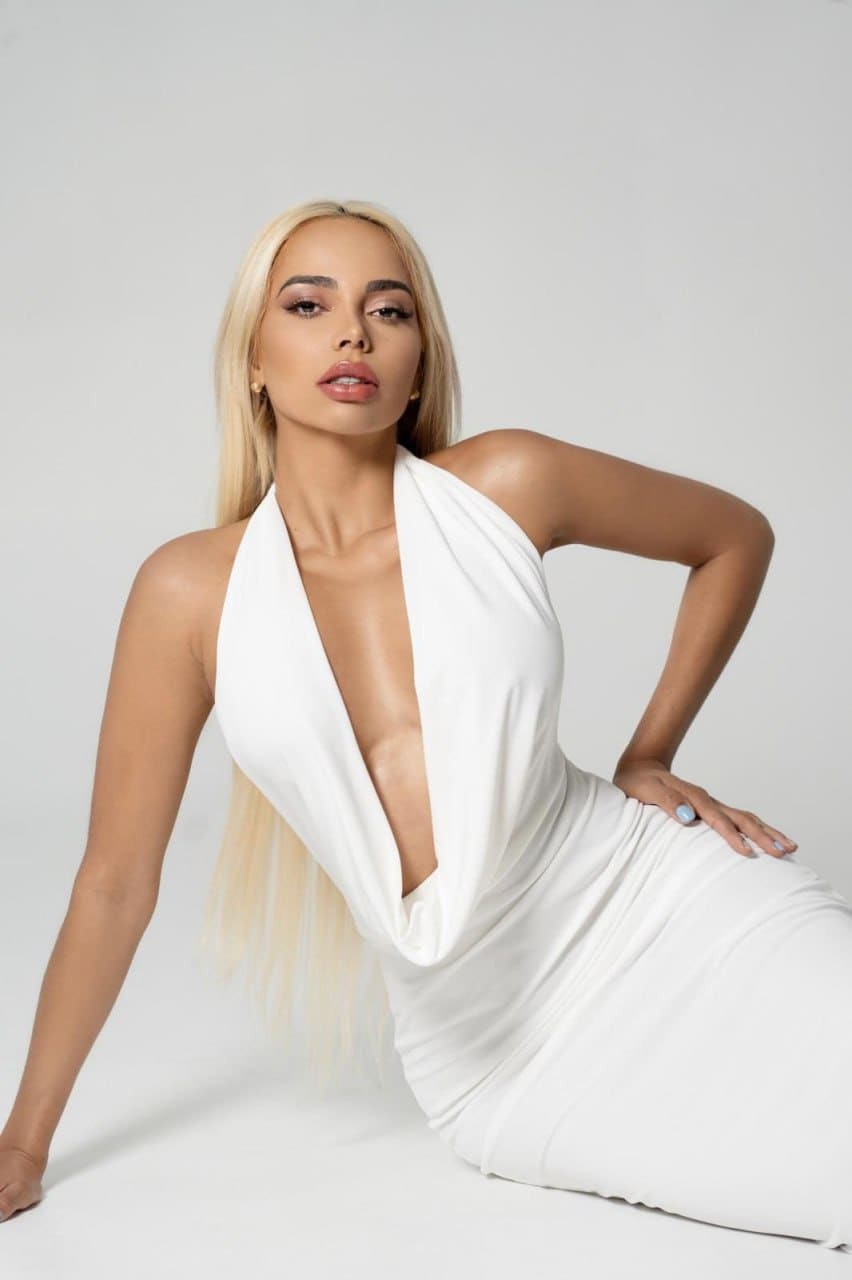 Valentina sits on a white surface in a white one-shoulder dress, left hand on hip, against a plain light background.