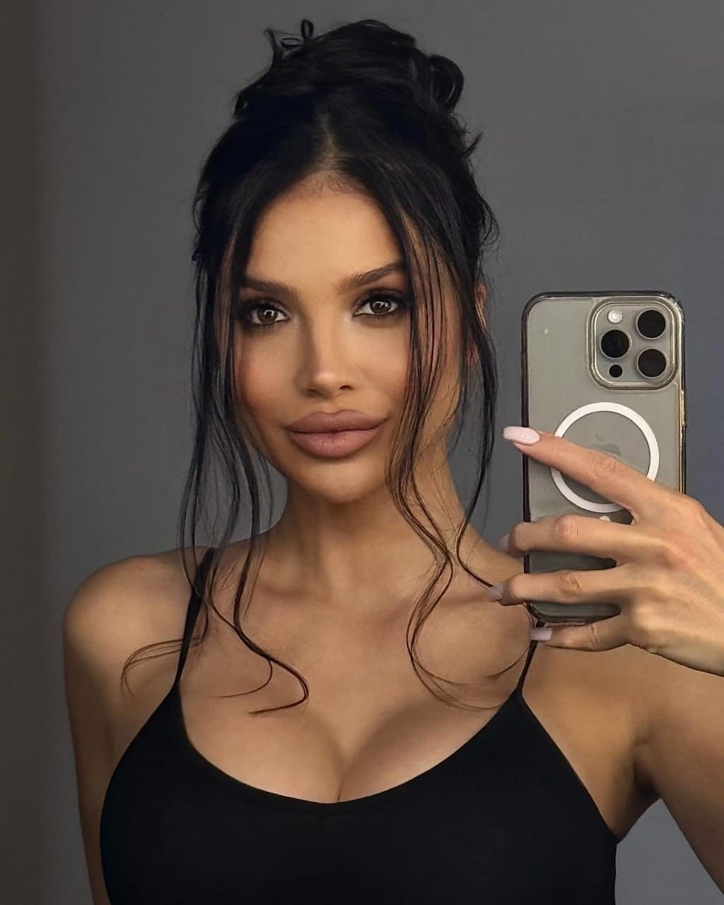 A woman with a messy updo takes a mirror selfie in a black spaghetti-strap top against a plain gray wall.