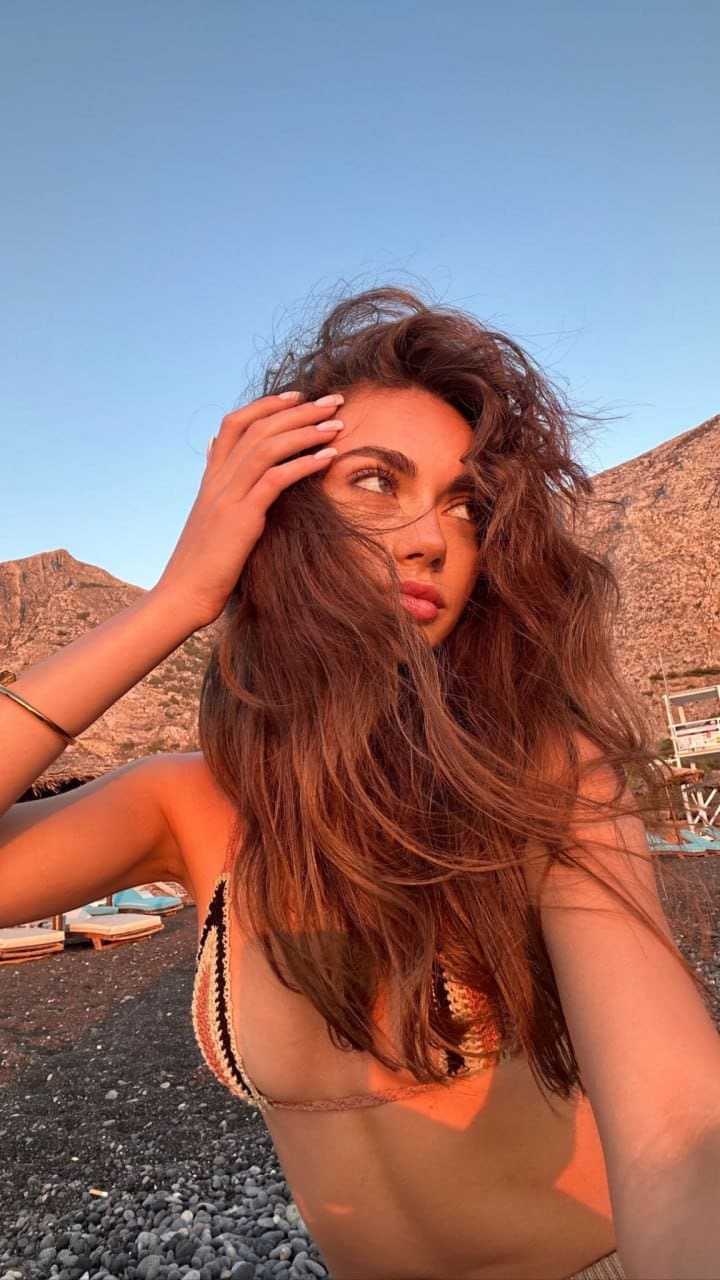 A woman in a patterned bikini top stands on a pebble beach, hand to forehead, looking away against rocky hills and sky.