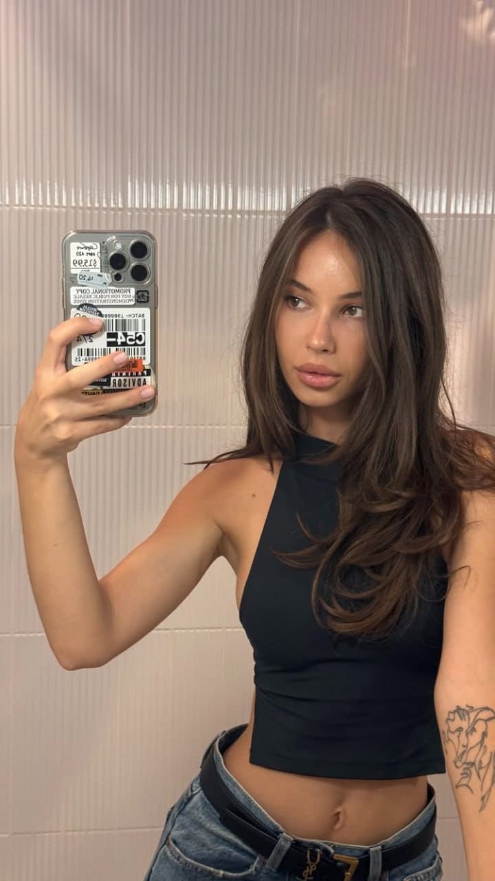 A mirror selfie shows a woman in a black crop top and blue jeans with a belt, standing before a ribbed wall.