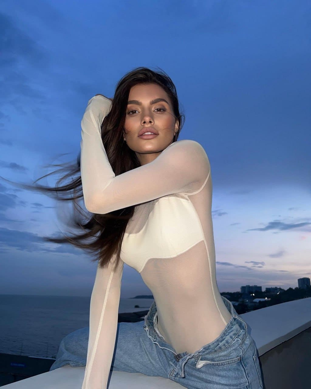A woman in a sheer top over a white bandeau and jeans sits on a rooftop ledge at dusk with a city skyline behind her.