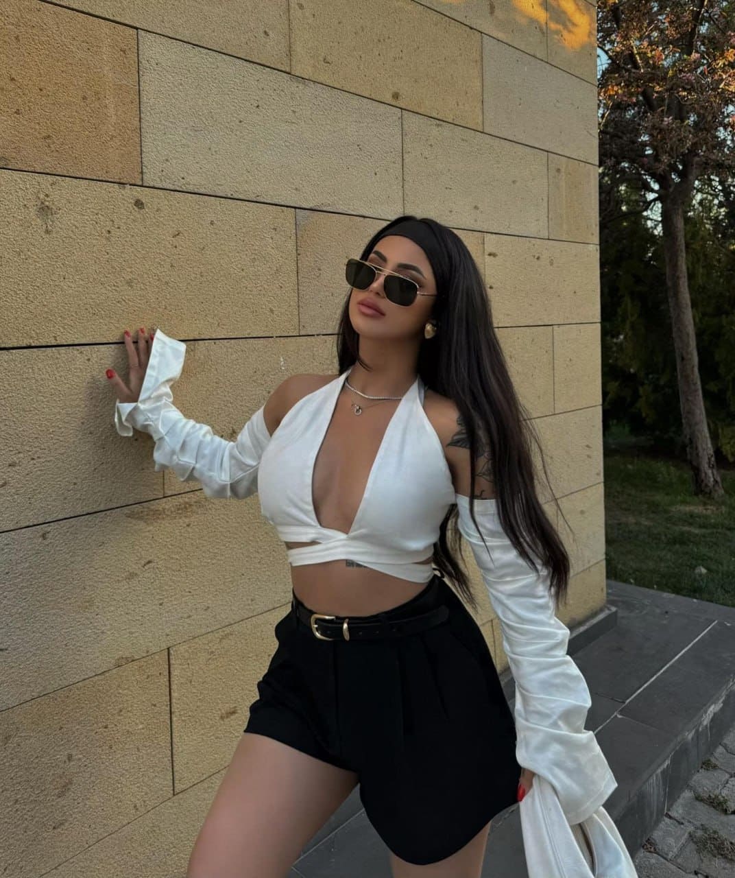 Leaning against a tan stone wall in a white halter crop top, black high-waisted shorts, and a draped white shirt, wearing sunglasses.