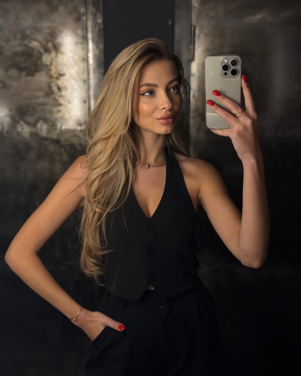 Mirror selfie in a dark room showing a black sleeveless top with a deep V-neck and trousers, hand on hip holding a glittery phone.
