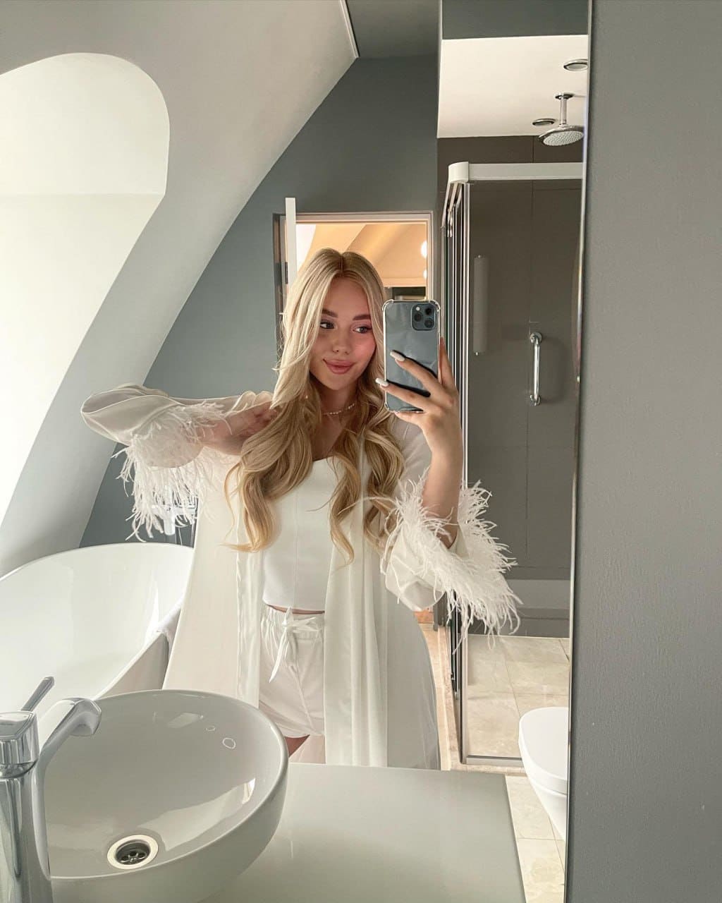 Mirror selfie of a woman in a white top, shorts, and feathered robe in a modern bathroom with sink and shower visible.