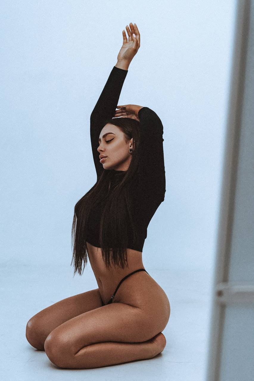 Kneeling on a light floor, she wears a black crop top and thong, one arm raised above her head.